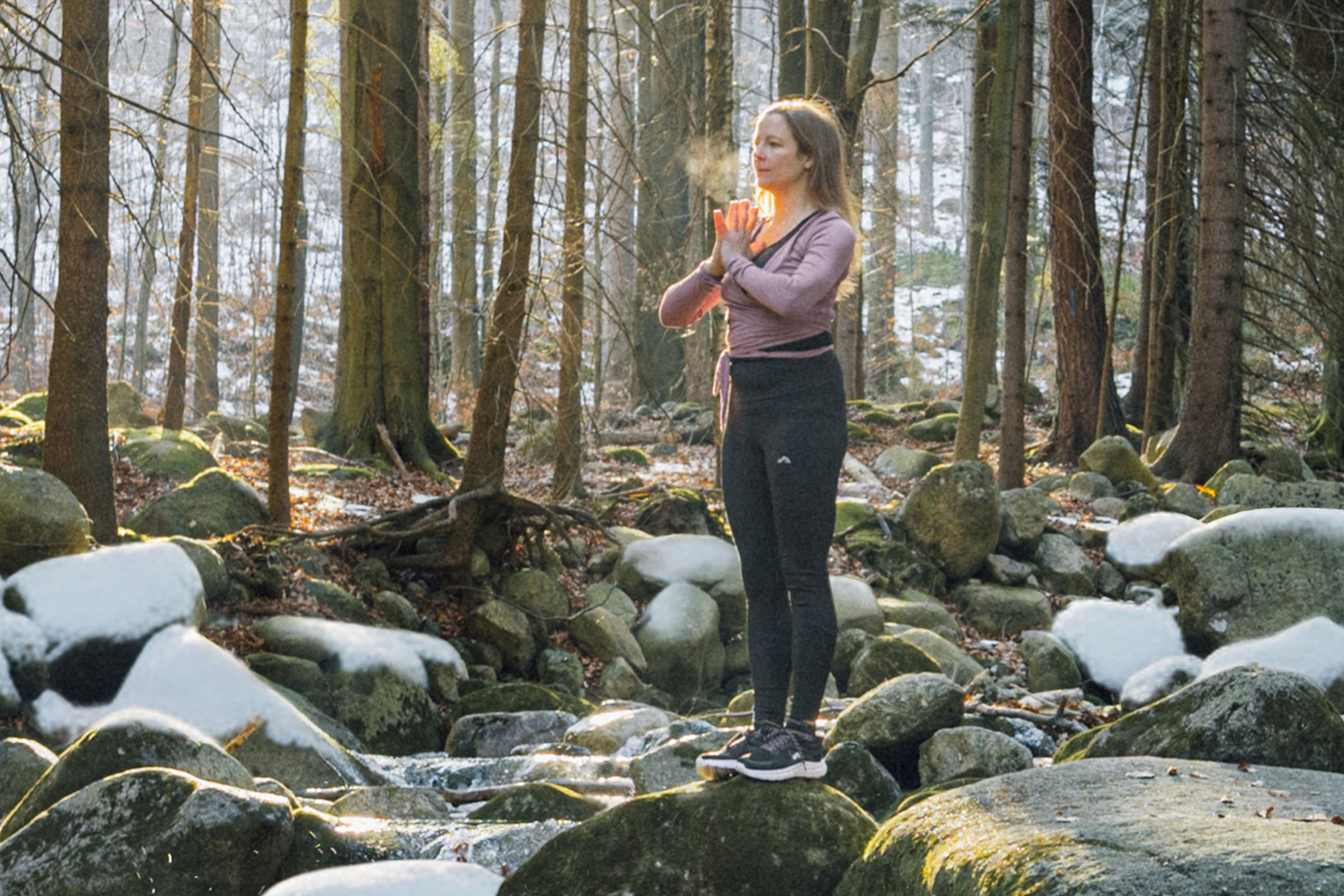 Vrouw in sportkleding die yoga beoefent in een winterbos bij een beekje.