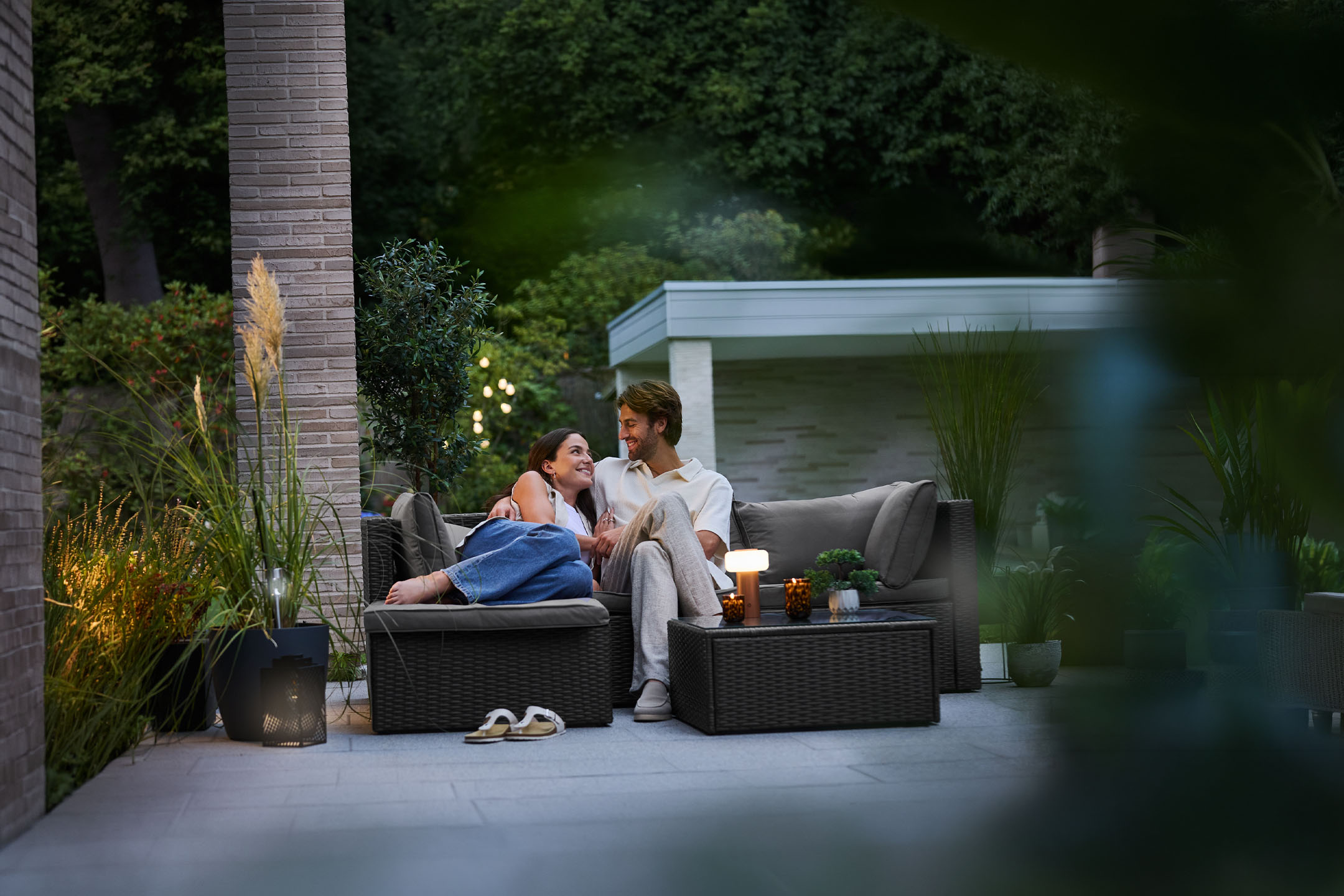 Koppel ontspant op een buitenbank op een terras met tuinmeubelen en verlichting.
