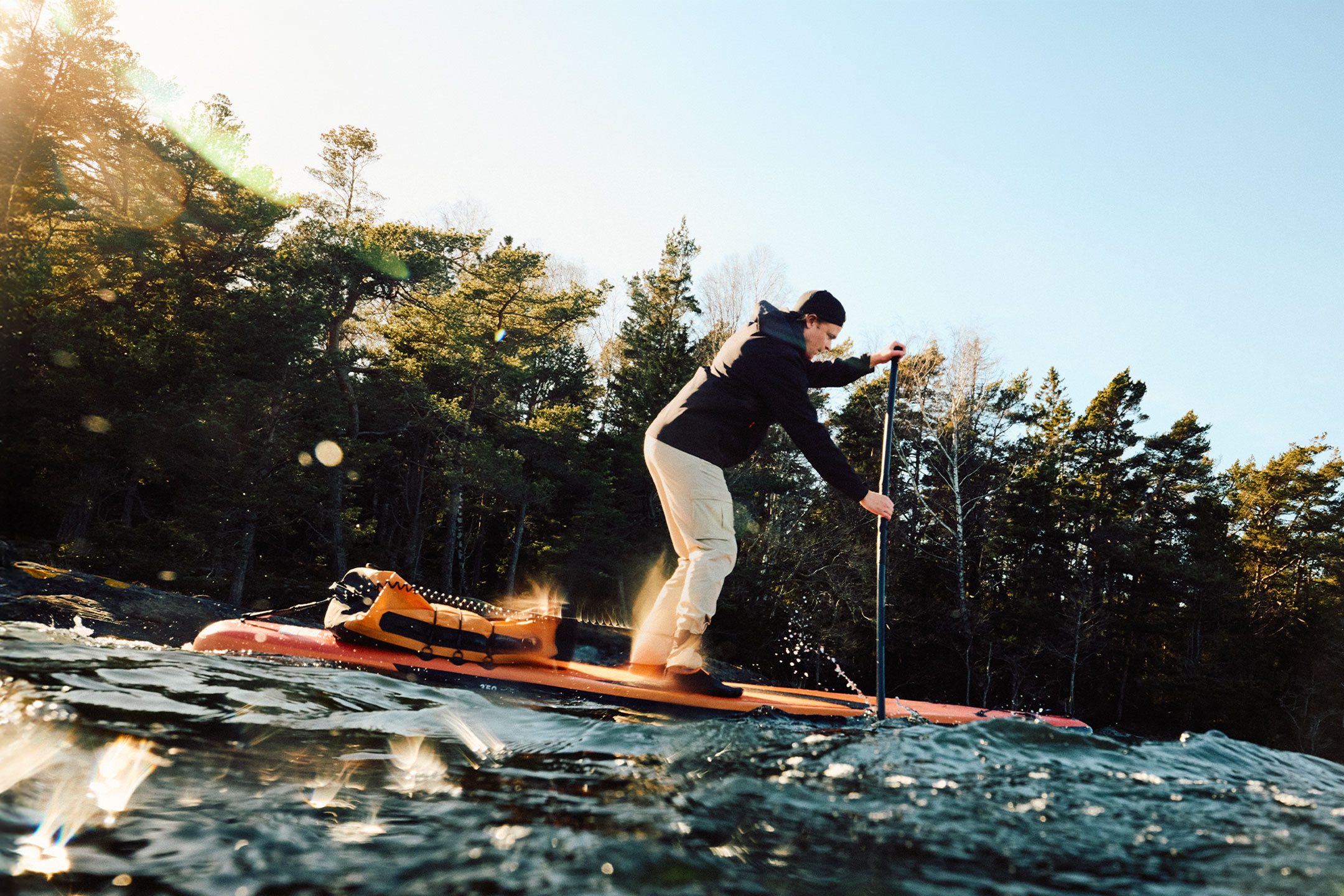 Man paddleboardt met een oranje drybag naast een bos.