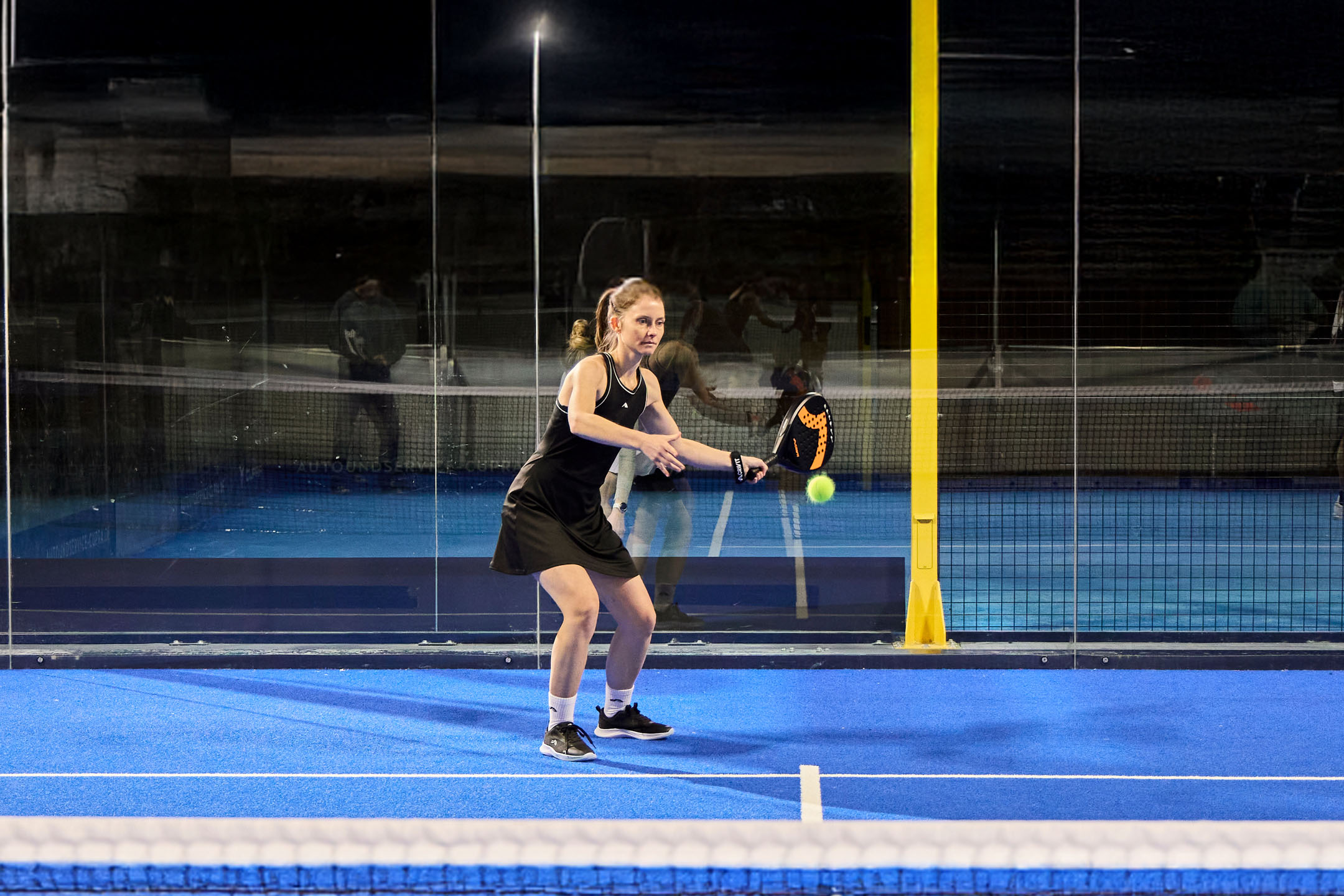 Vrouw in sportkleding speelt padel op een blauwe baan met racket en bal.