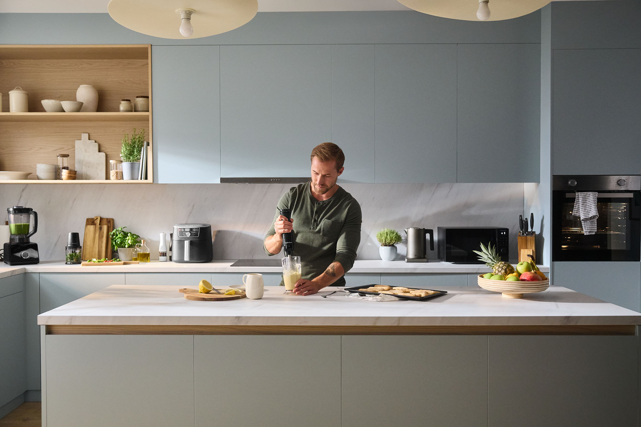 Man gebruikt staafmixer in moderne keuken met apparaten en fruit.
