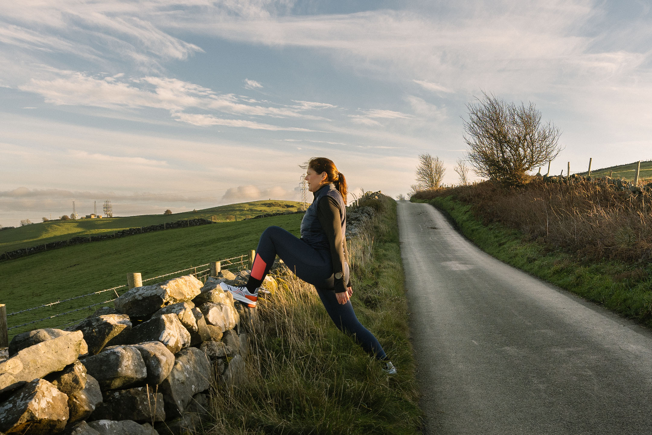 Vrouw in sportkleding rekt zich uit op een stenen muur langs een landweg.