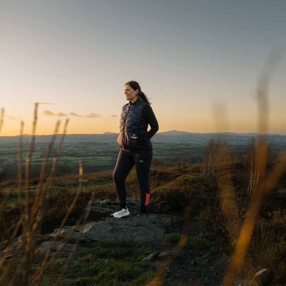 Vrouw in gewatteerd vest en legging staat op een heuveltop bij zonsondergang.