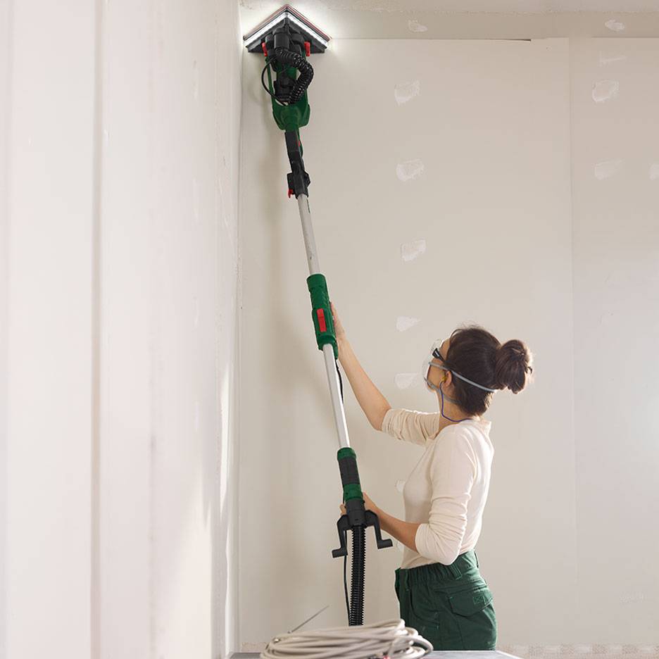 Vrouw schuurt een muur met een telescopische wandschuurmachine in een hoek.