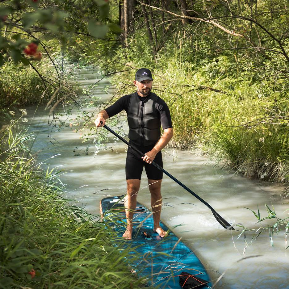 Man in wetsuit en zwemvest staat op een paddleboard in een beekje.