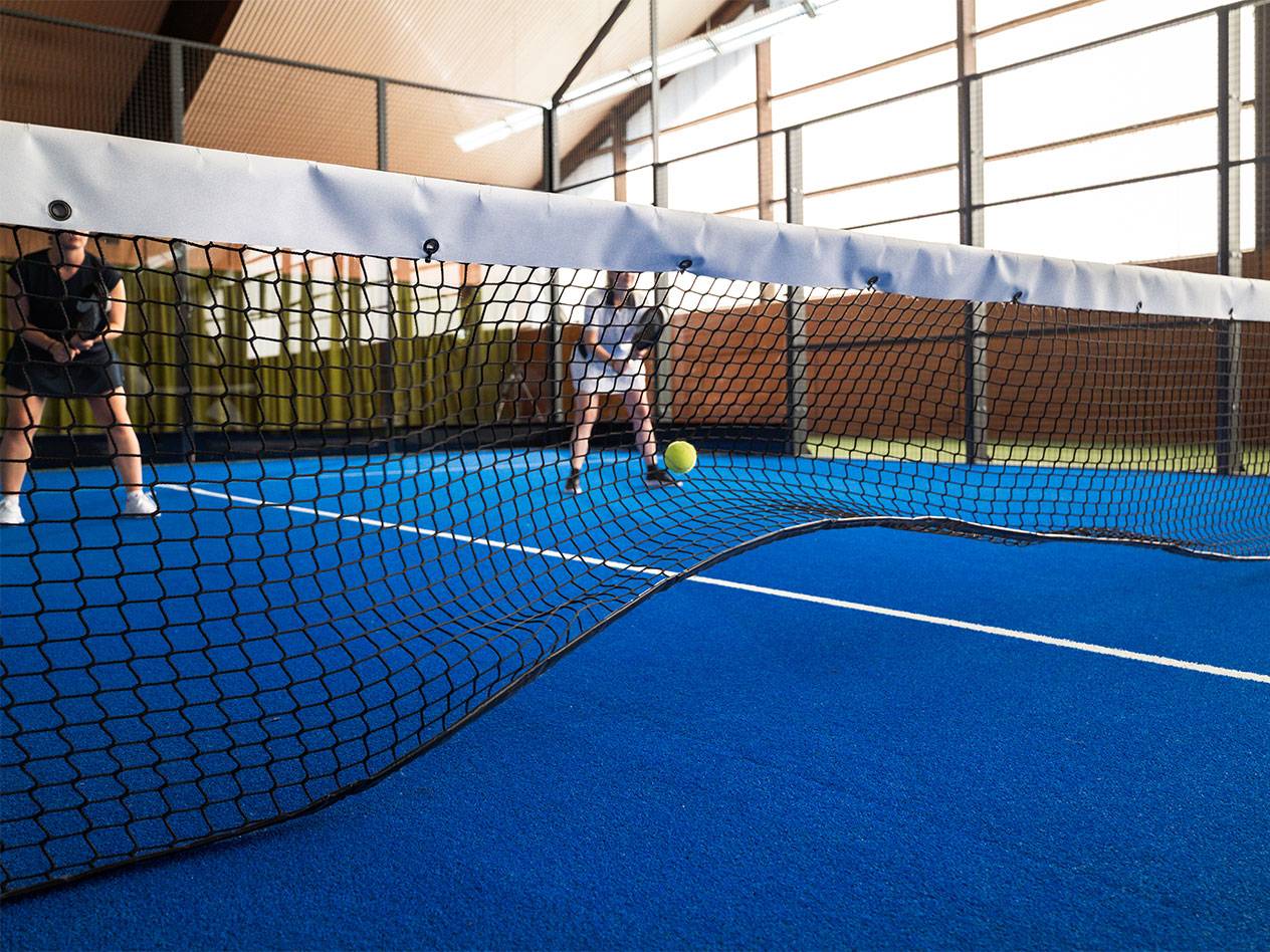 Twee vrouwen spelen padel op een blauw veld, met het net op de voorgrond.