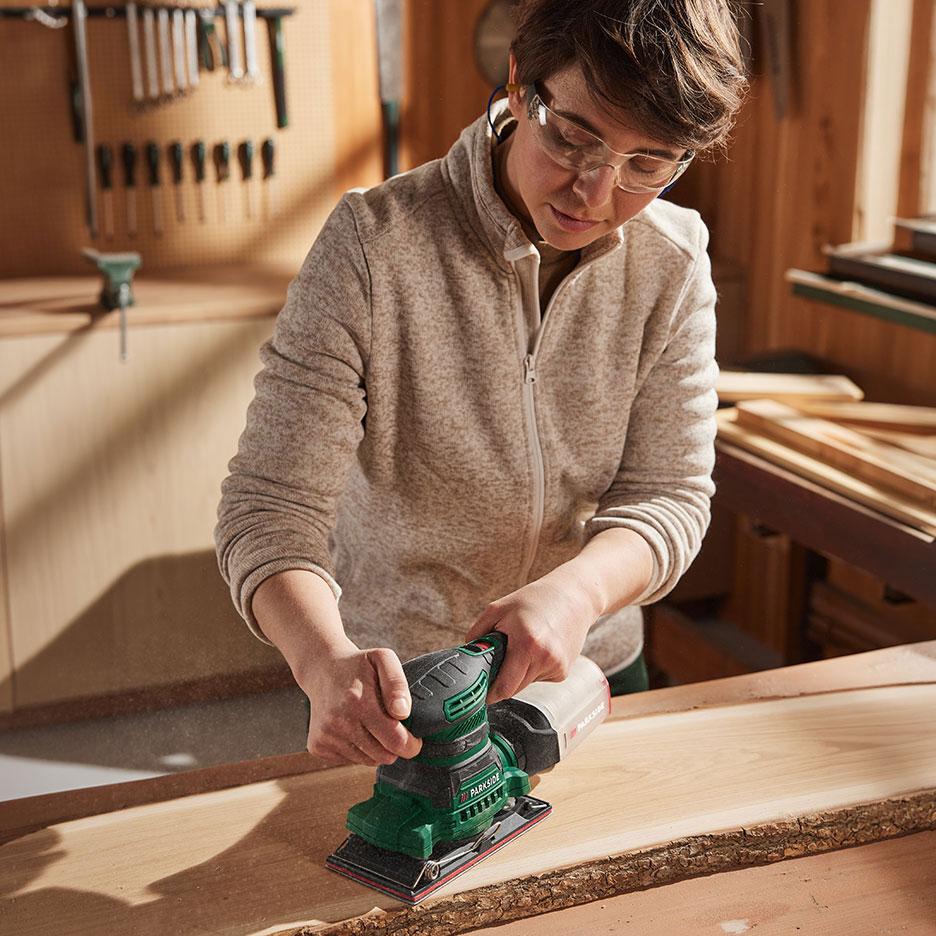 Vrouw met elektrische schuurmachine schuurt een stuk hout in een werkplaats.