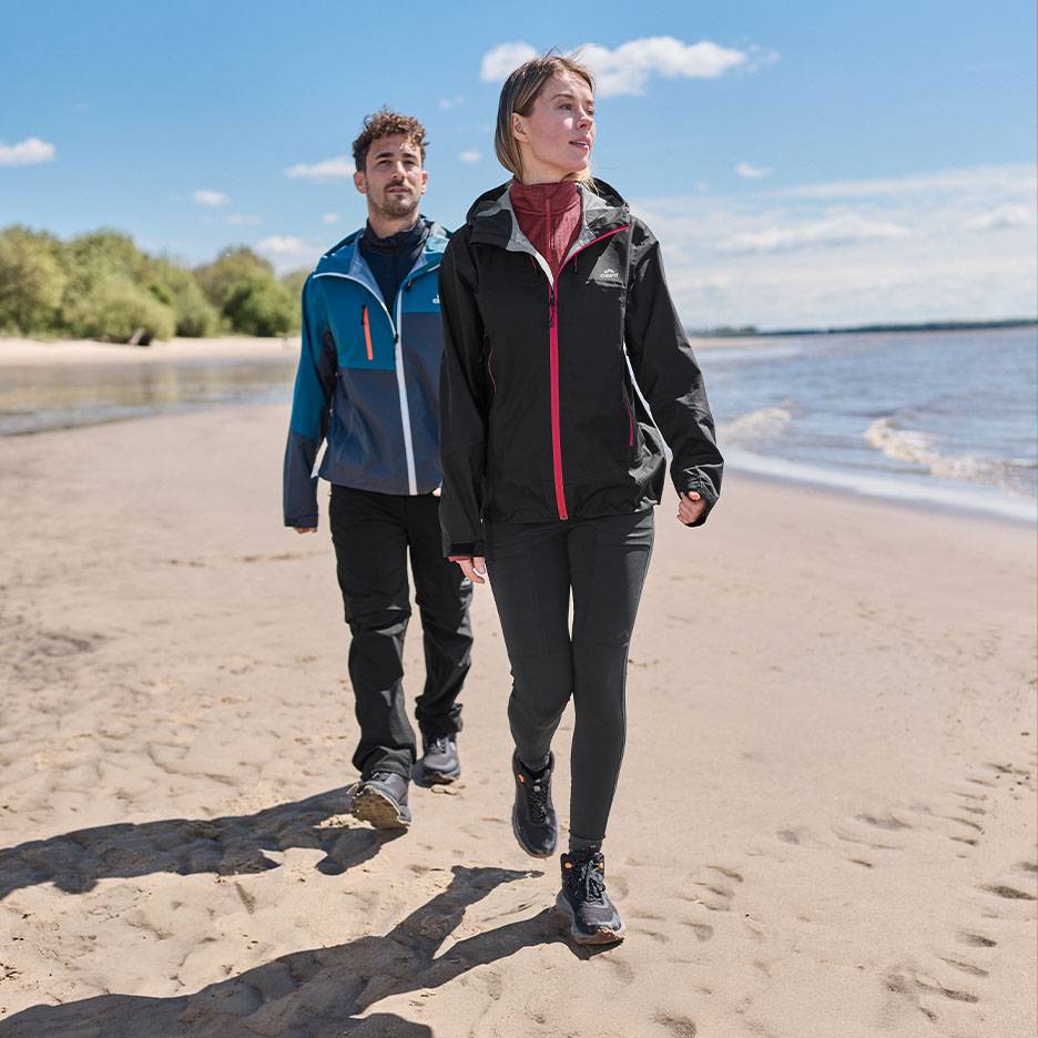 Koppel in wandeljassen en -broeken wandelt op een zandstrand aan zee.