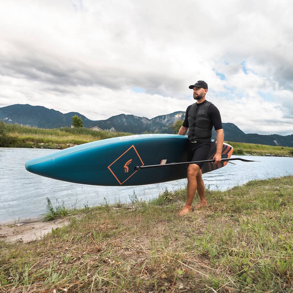 Man met zwemvest en pet draagt een paddleboard en peddel langs een rivier.