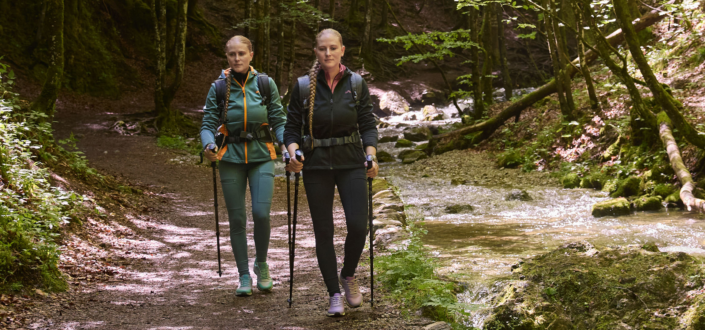 Twee vrouwen wandelen langs een beek in een bos met wandelstokken.