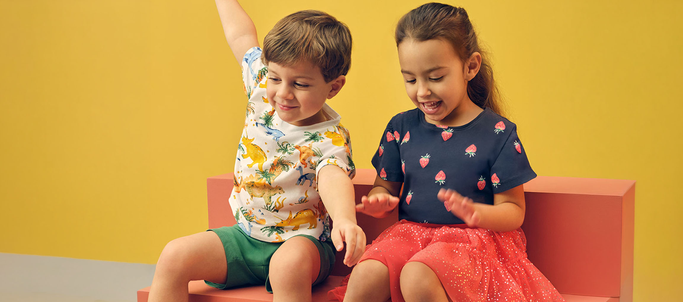 Twee kinderen, een jongen en een meisje, zitten op een roze bankje in zomerkleding.