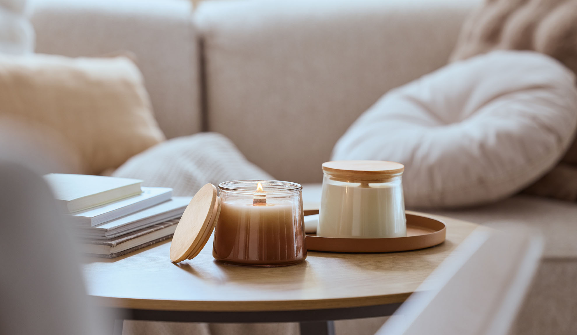 Twee geurkaarsen op een houten tafel, één brandend, met een gezellige bank op de achtergrond.