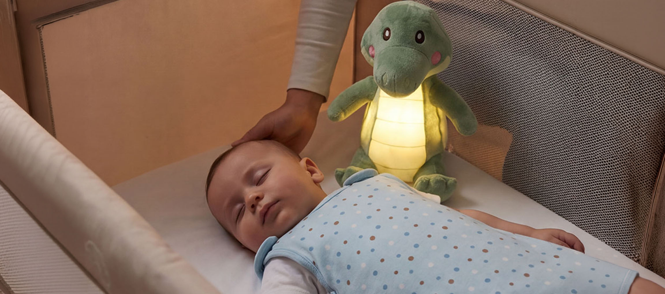Baby slaapt in wieg met knuffel-nachtlampje, volwassene aait het hoofd van de baby.