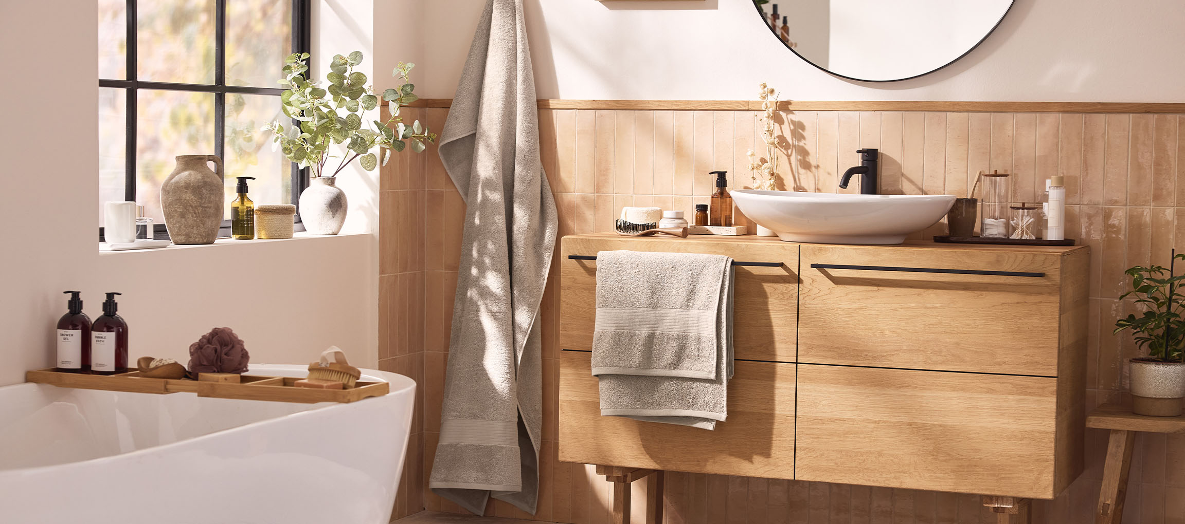 Een moderne badkamer met een houten badmeubel, witte wastafel, ronde spiegel en badkuip met badaccessoires.