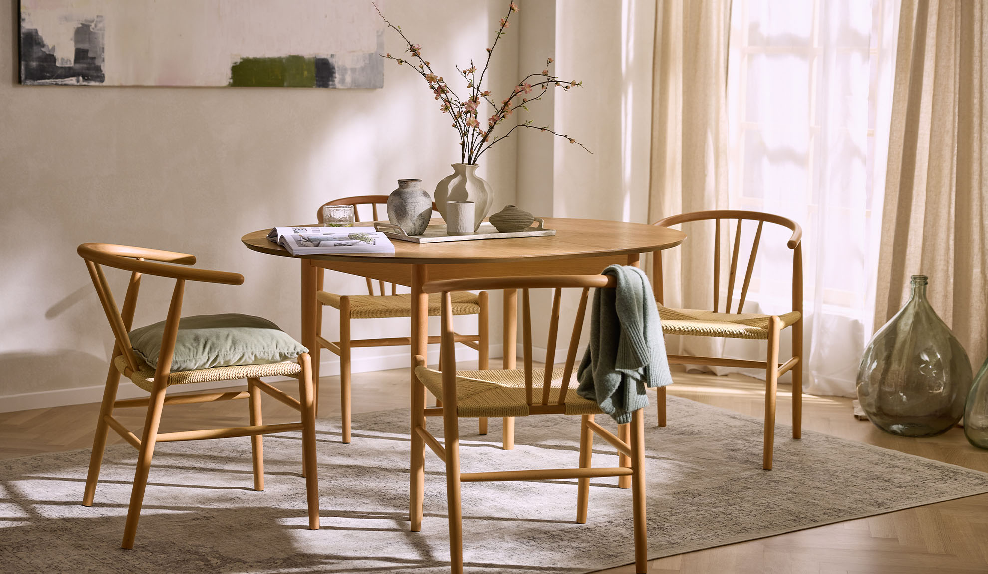 Lichte eetruimte met ronde houten tafel, houten stoelen met touwzittingen en bloemdecoratie.
