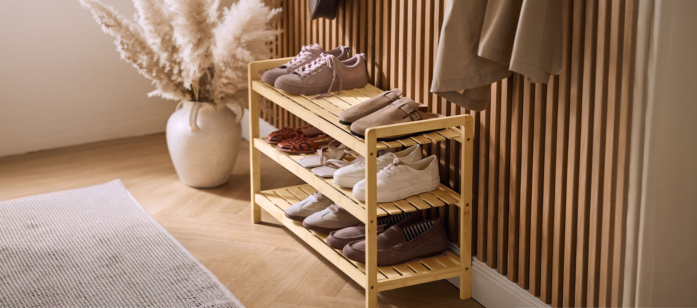 Houten schoenenrek met diverse schoenen, een vaas met pampasgras en een jas aan de muur.