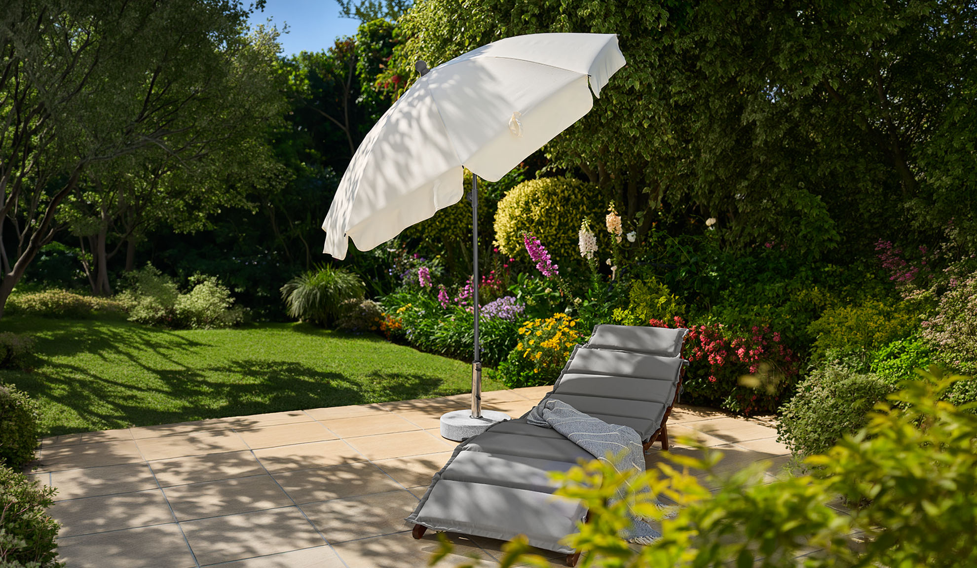 Grijze ligstoel met witte parasol op een zonnig terras met weelderige tuin.