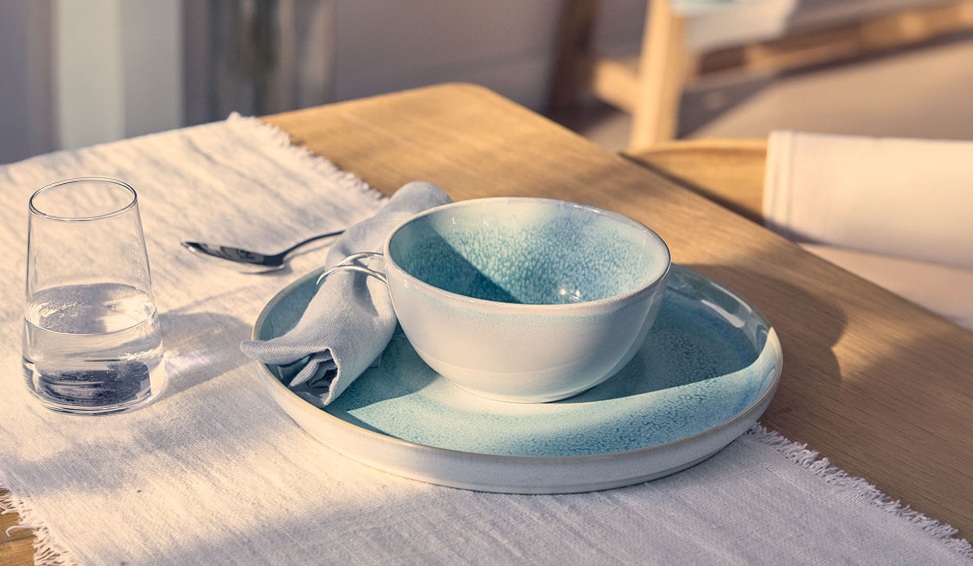 Gedekte tafel met blauwe kom, bord, glas water en servet op een houten tafel.