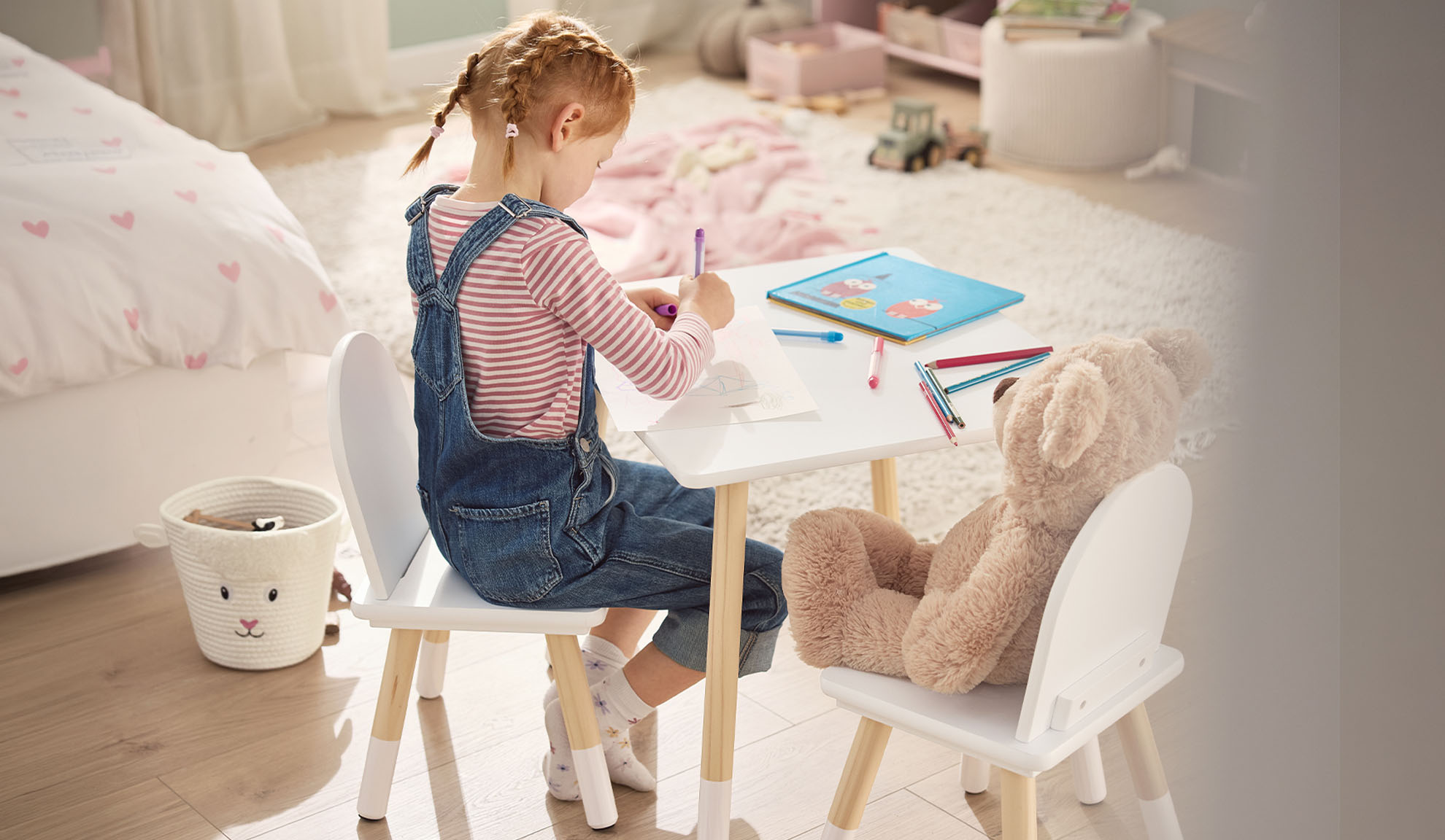 Meisje tekent aan een witte tafel met een teddybeer, in een kinderkamer.