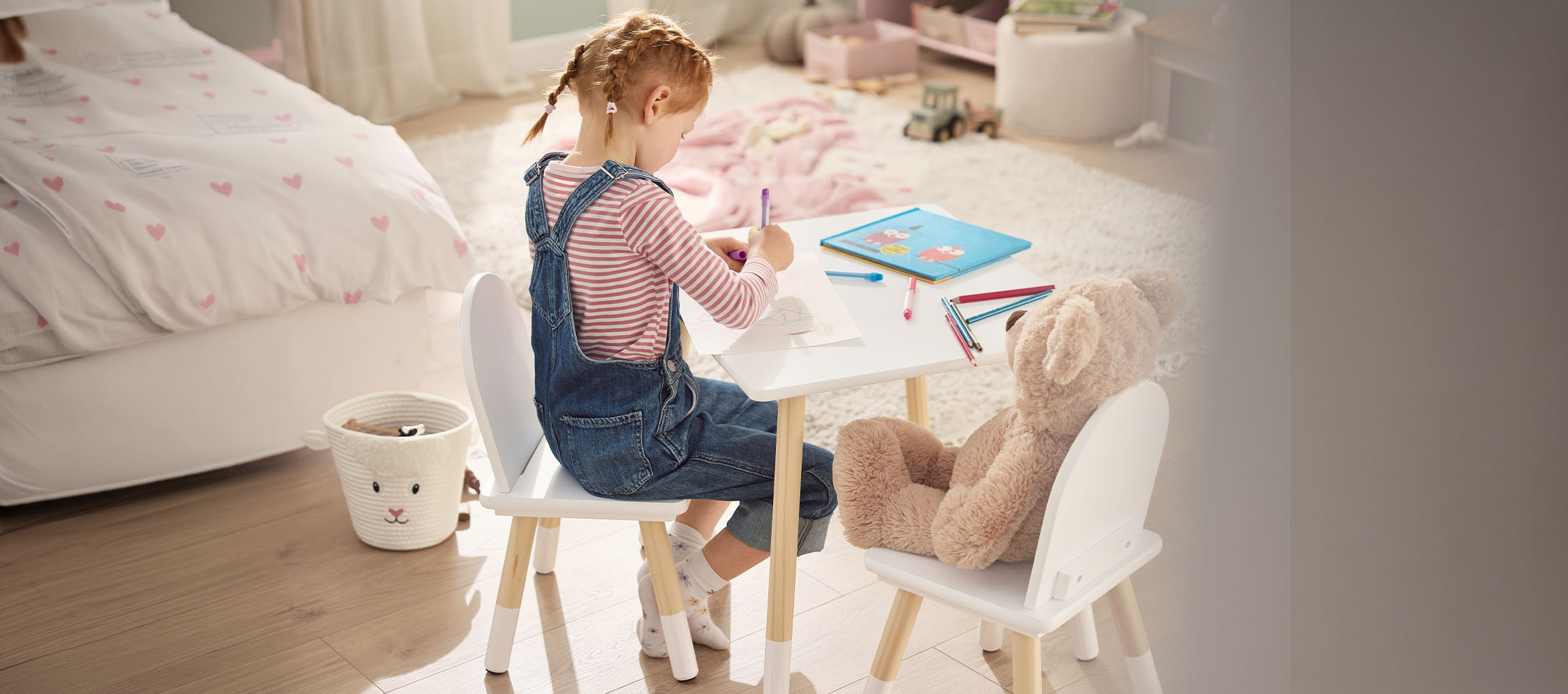 Meisje tekent aan een witte tafel met stoelen, een teddybeer naast haar.