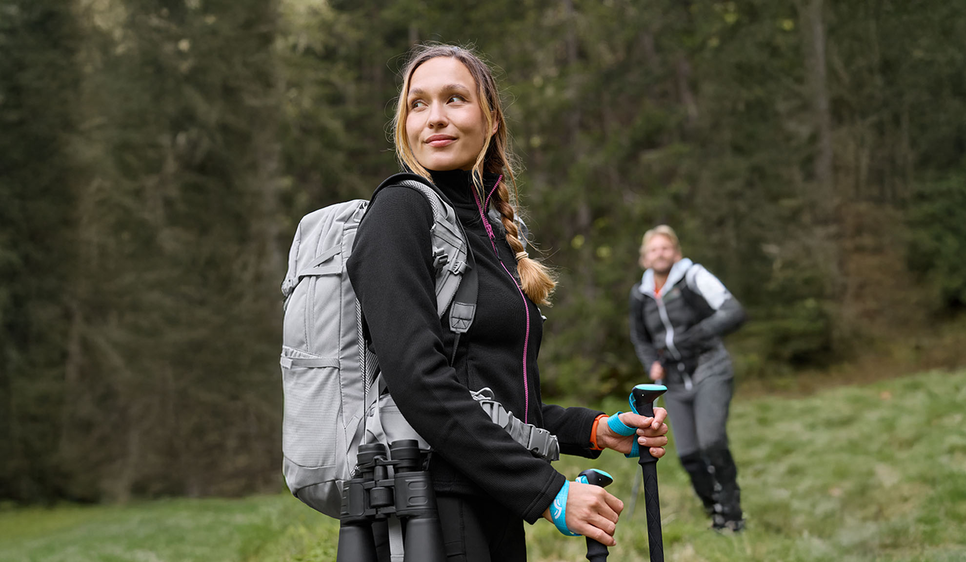 Vrouw met wandelrugzak en trekkingstokken, man op de achtergrond.