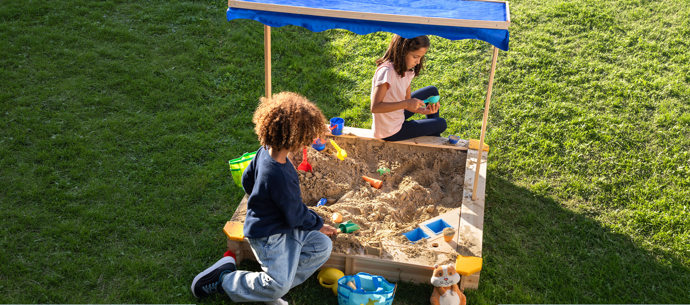 Twee kinderen spelen in een houten zandbak met een blauw afdak, omringd door groen gras.