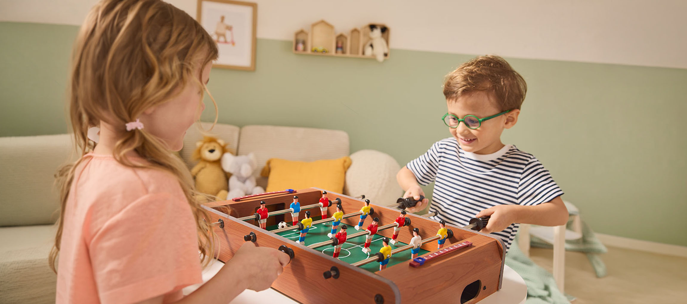 Twee kinderen spelen tafelvoetbal, een meisje in een roze shirt en een jongen in een gestreept shirt.