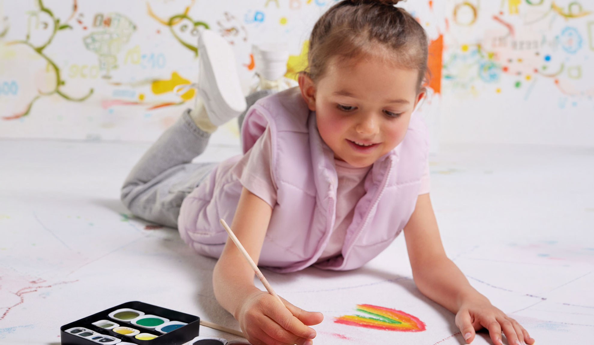 Een kind schildert een regenboog op papier met waterverf, in een roze vest.