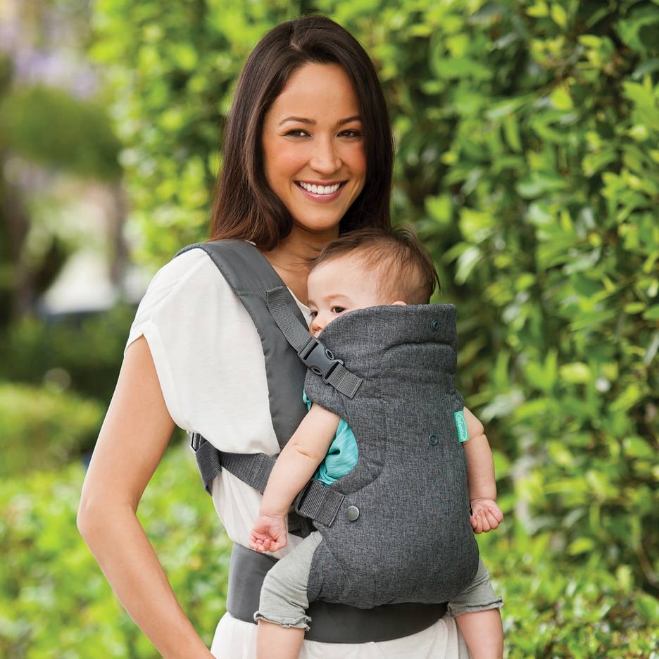 Lachende vrouw in een grijze draagzak met een baby buiten.