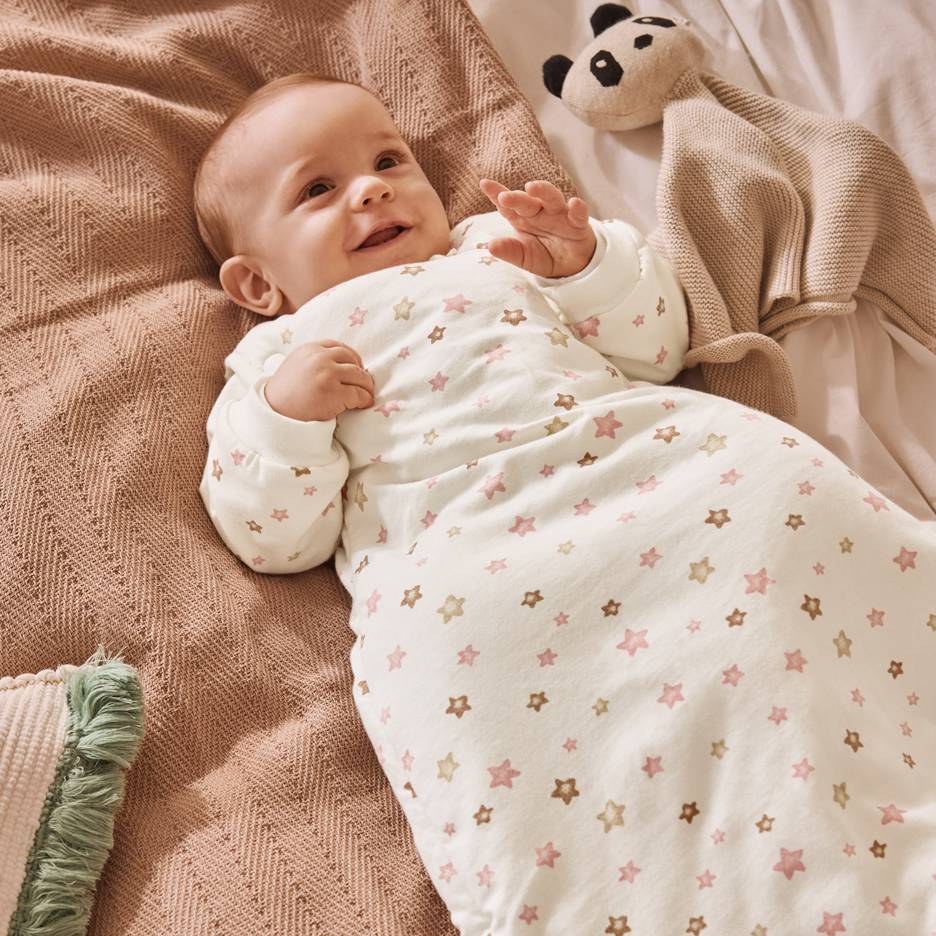 Lachende baby in witte slaapzak met roze en bruine sterren, liggend op een deken.