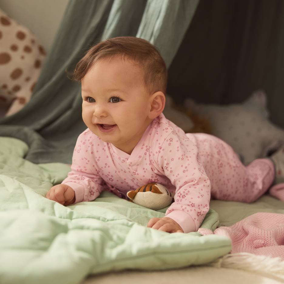 Baby in roze sterrenpyjama, liggend op een groene deken met een knuffel.
