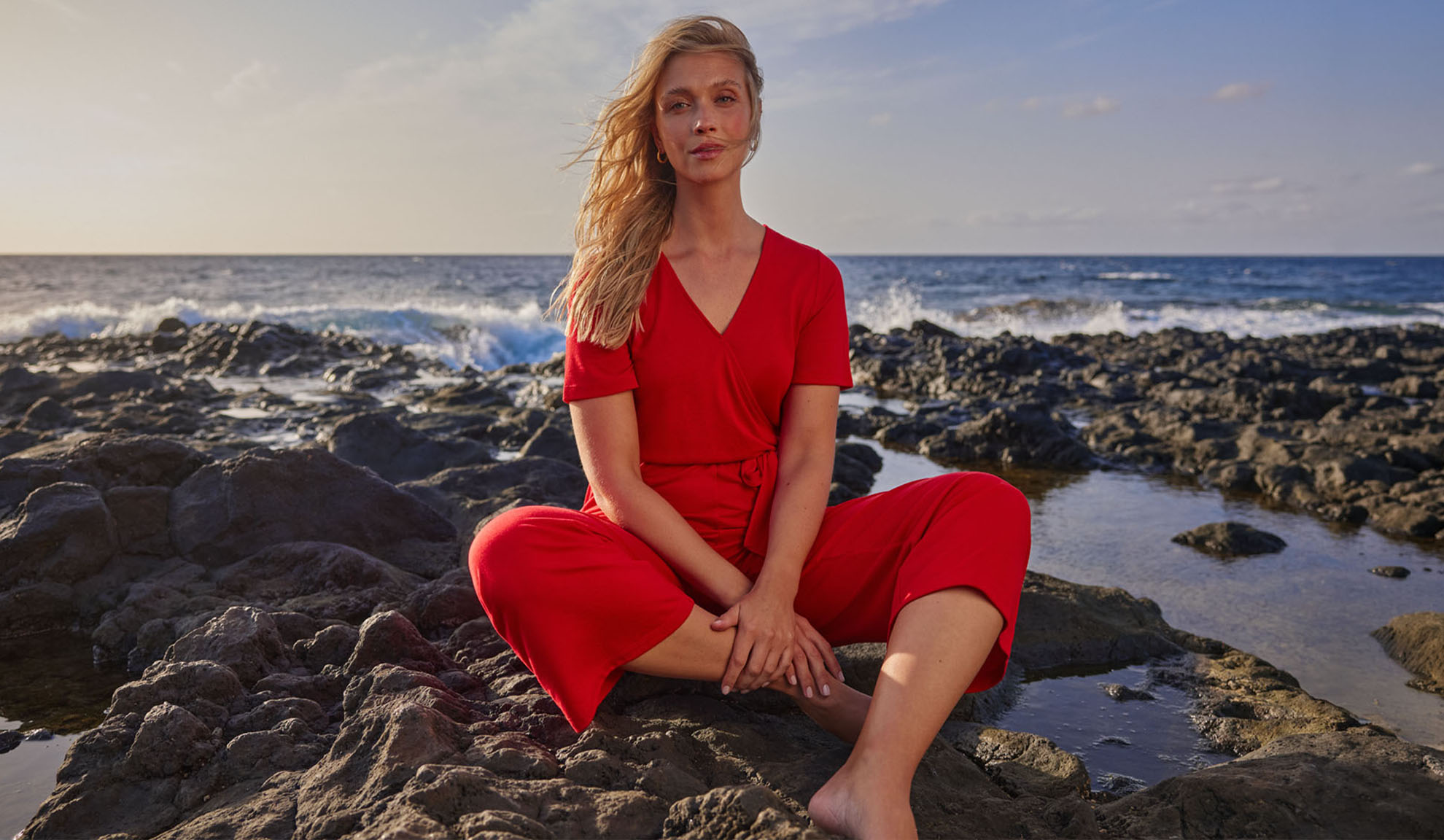 Vrouw in rode jumpsuit zit op rotsen bij de zee.