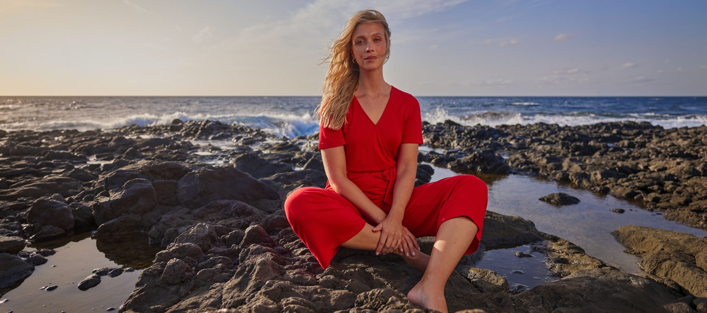 Vrouw in rode jumpsuit zit op een rotsachtig strand bij de zee.