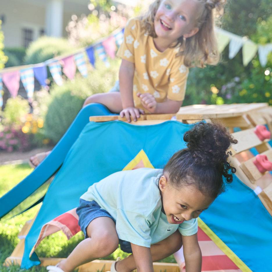 Twee blije kinderen spelen op een houten klimrek met een blauwe tent in een zonnige tuin.