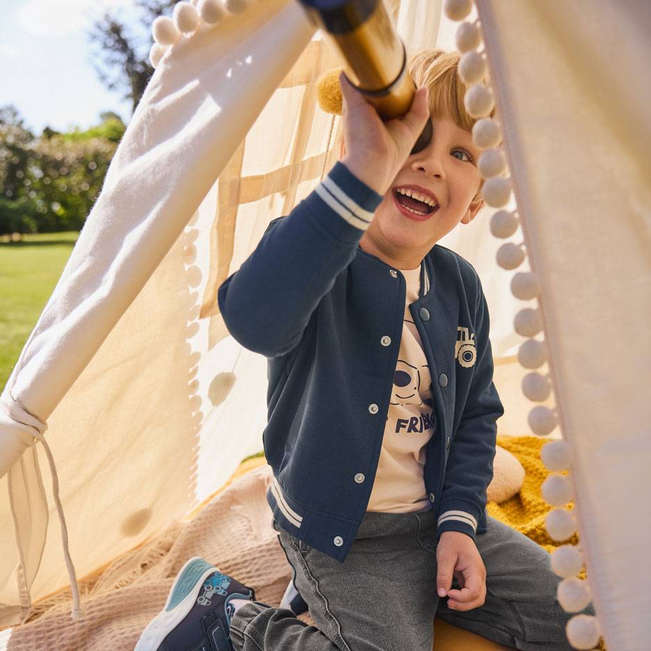 Lachende jongen in blauw jasje en beige T-shirt, kijkend door een telescoop in een tent.