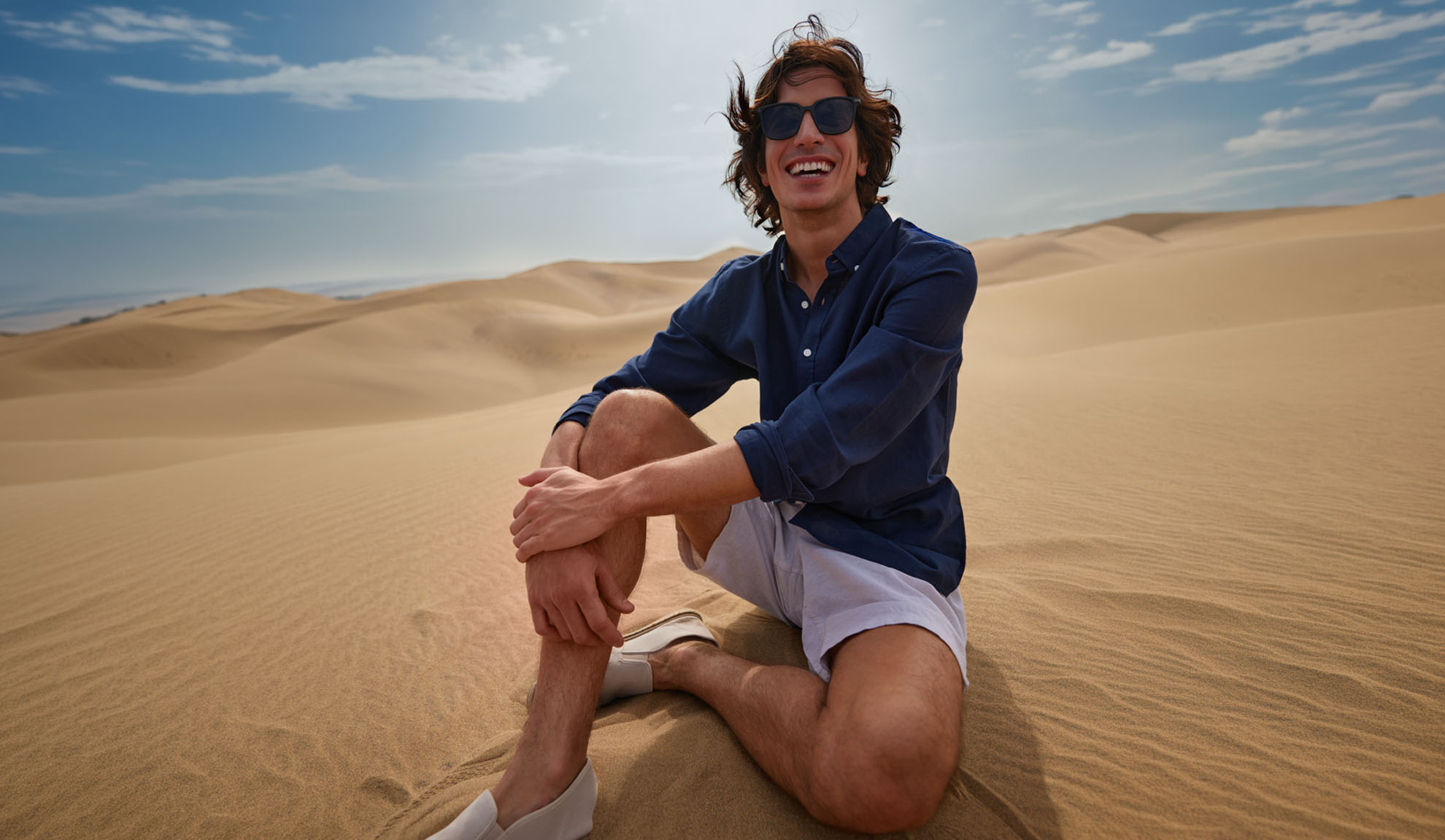 Man in donkerblauw overhemd en lichte shorts zit lachend op zandduinen.