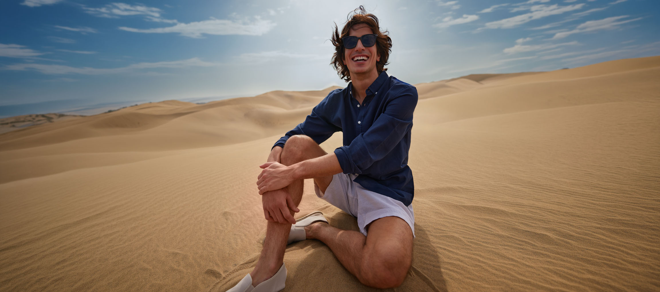 Man in donkerblauw overhemd en korte broek zit lachend in de zandduinen.