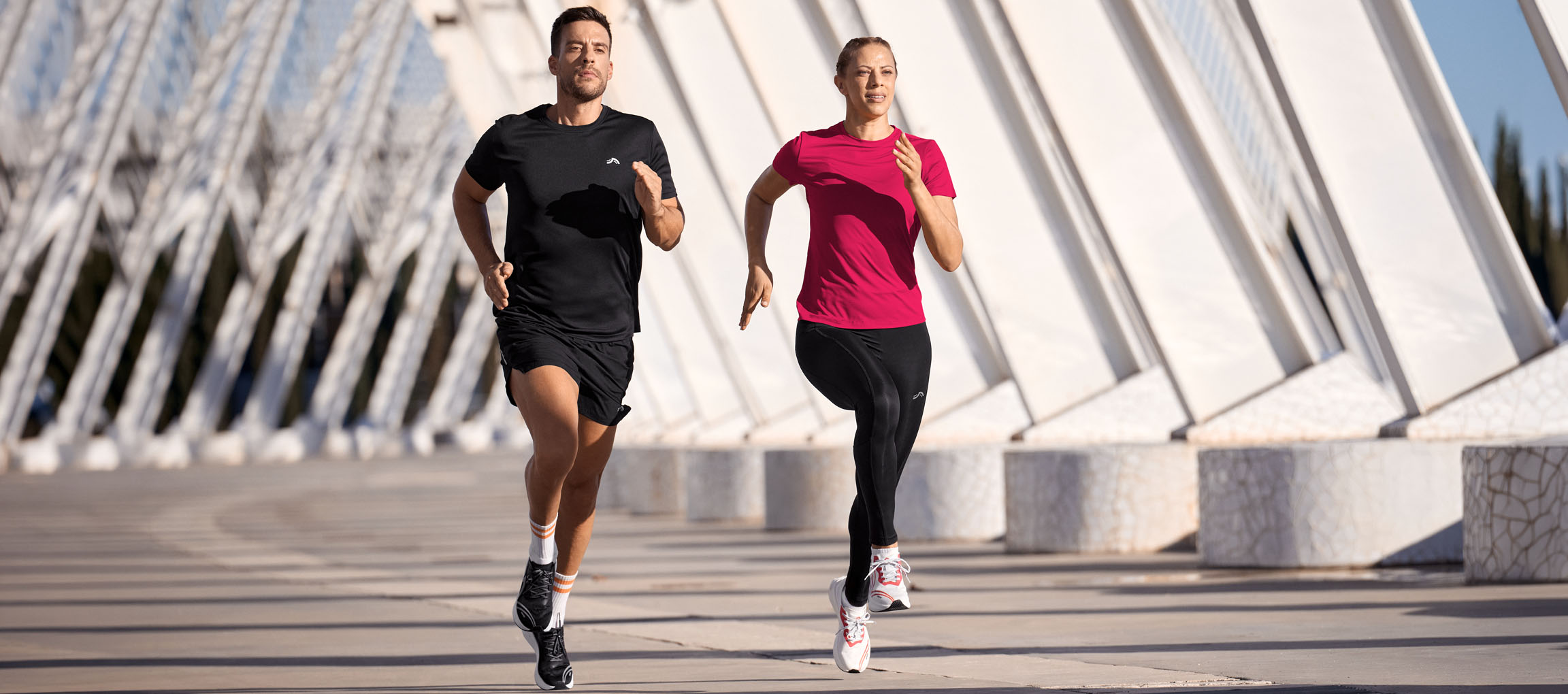 Man en vrouw rennen in sportkleding, zwarte en roze tops, zwarte shorts en leggings.