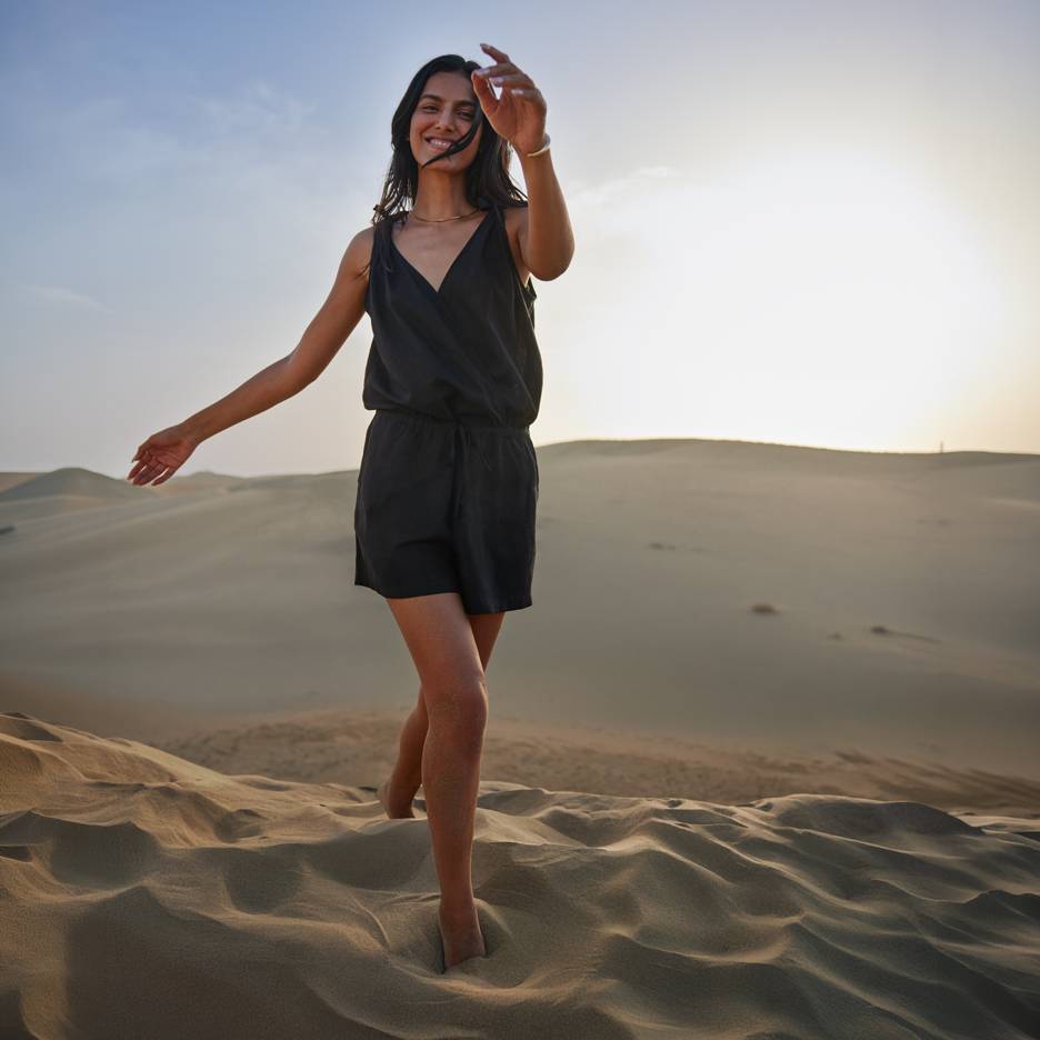 Vrouw in zwarte jumpsuit op blote voeten in zandduinen bij zonsondergang.