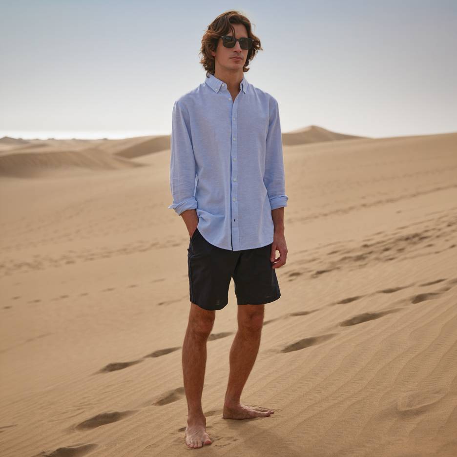 Man in blauw overhemd en zwarte korte broek, blootsvoets staand op zandduinen.