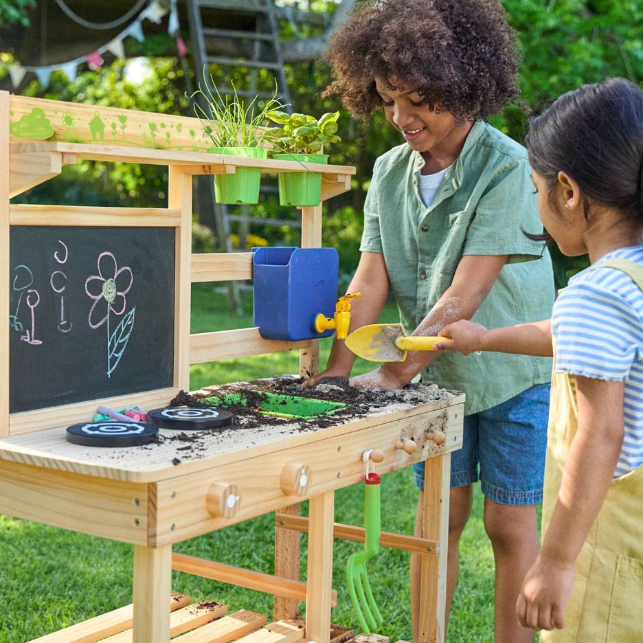 Twee kinderen spelen met een houten modderkeuken, met een krijtbord en potplanten.