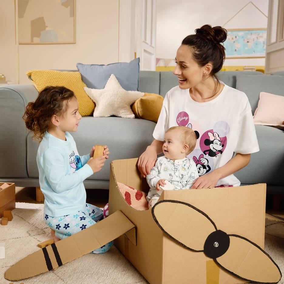 Moeder en kinderen spelen met een kartonnen doos, waarbij moeder een T-shirt met stripfiguren draagt.