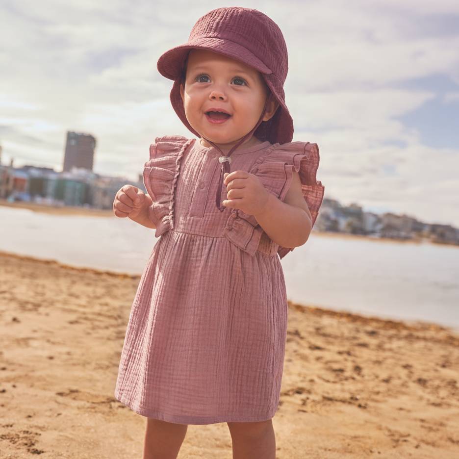 Baby in roze jurk en hoed op het strand.