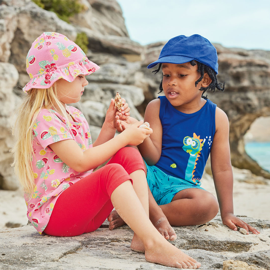 Twee kinderen op een strand, één met een roze zonnehoed en fruitpatroon top, de ander met een blauwe pet en dinosaurus tanktop, schelpen vasthoudend.