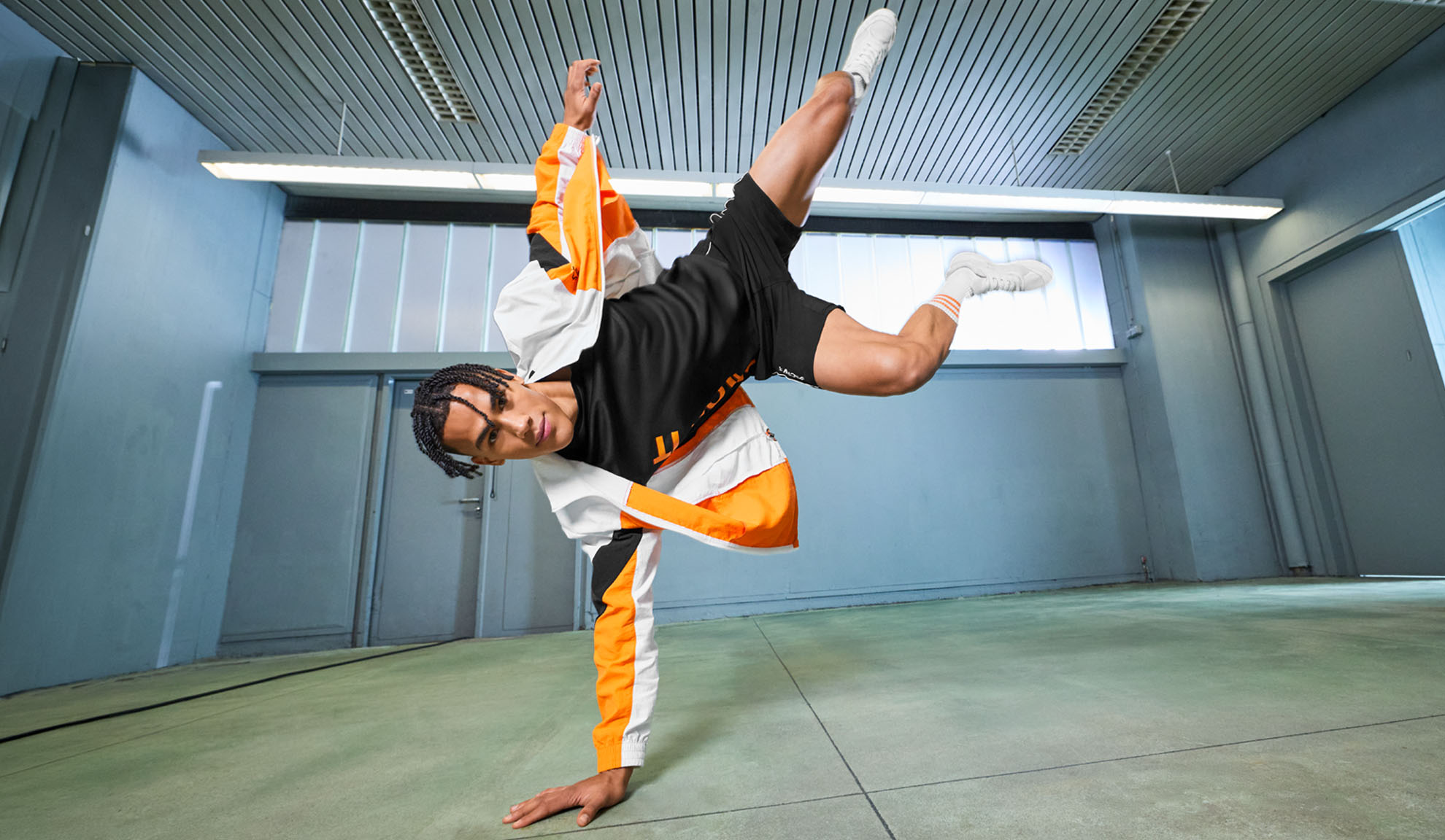 Man in sportkleding, zwart T-shirt, korte broek en oranje-witte jas, voert een dansbeweging uit.