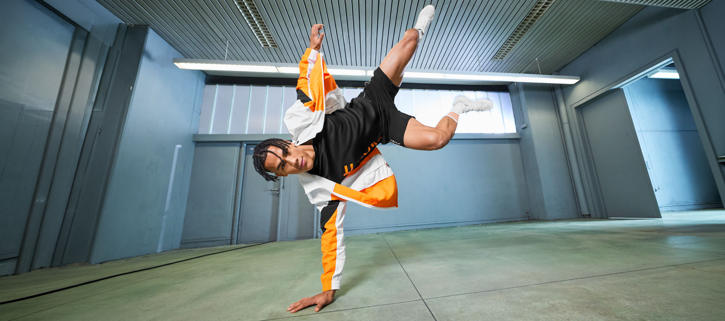 Man in sportkleding, zwart T-shirt en oranje-witte jas, danst in een kamer.