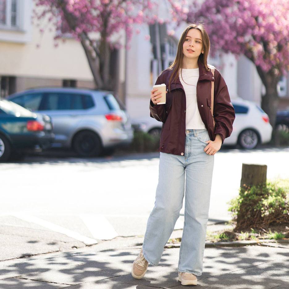 Vrouw in jas en jeans met koffie op straat met bloeiende bomen.