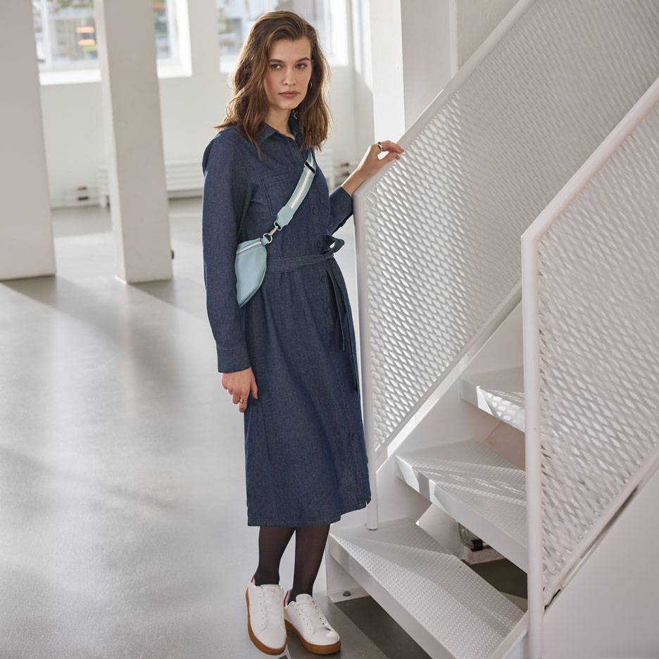 Vrouw in een denim overhemdjurk, zwarte panty en witte sneakers, met een lichtblauwe heuptas.