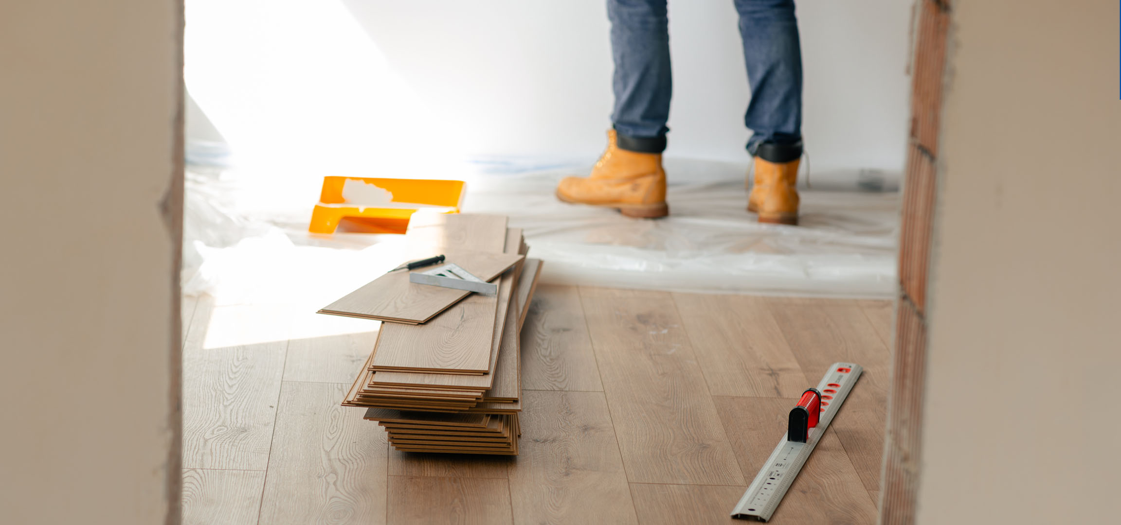 Laminaatvloerplanken en installatiegereedschap op een houten vloer, met een persoon in laarzen.