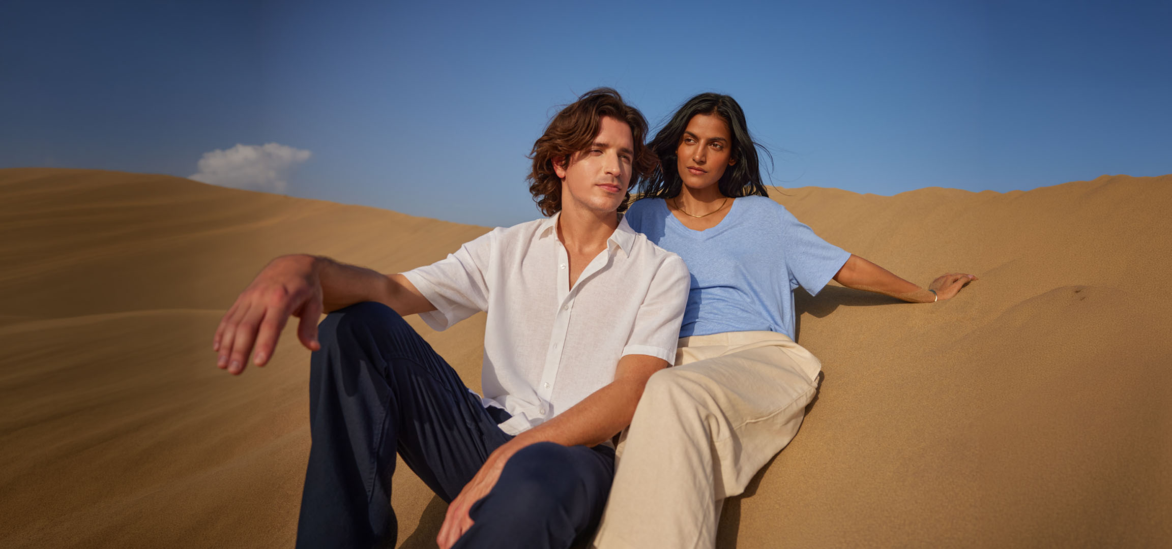 Man en vrouw zitten in zandduinen, gekleed in linnen overhemden en broeken.
